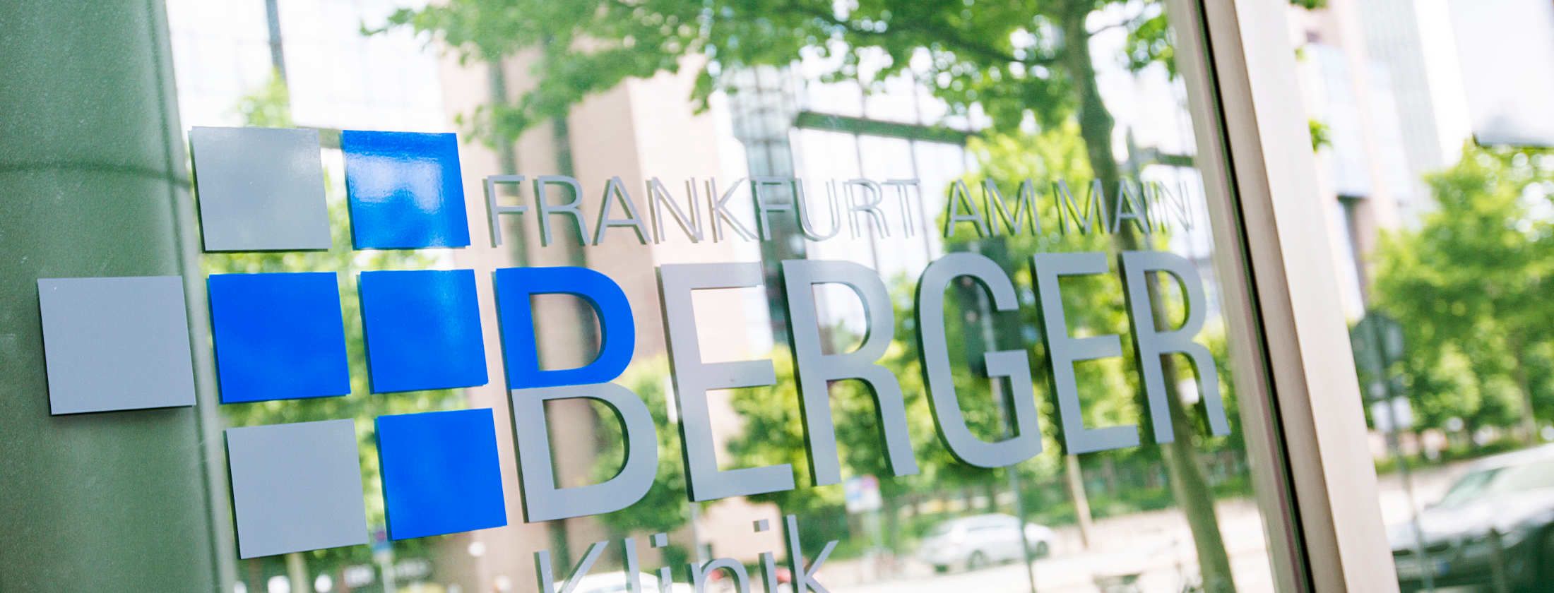 Aufnahme des Firmenlogos der privaten Berger Klinik auf der Mainzer Landstraße. Das Firmensymbol ist als Folienplott auf einer Außenscheibe angebracht. In den mächtigen Fenstern des Operationszentrums spiegeln sich grüne Bäume und weitere Gebäude.