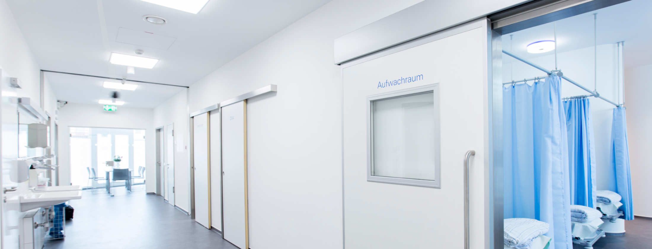 Ein langer heller und lichtdurchfluteter Flur des Operationszentrum Berger Klinik im Frankfurter Westend. Eine Schiebetür ist offen und lässt den Blick in den Aufwachraum nach Operationen zu. Der Aufwachraum bietet Platz für gleichzeitig 6 Patienten.