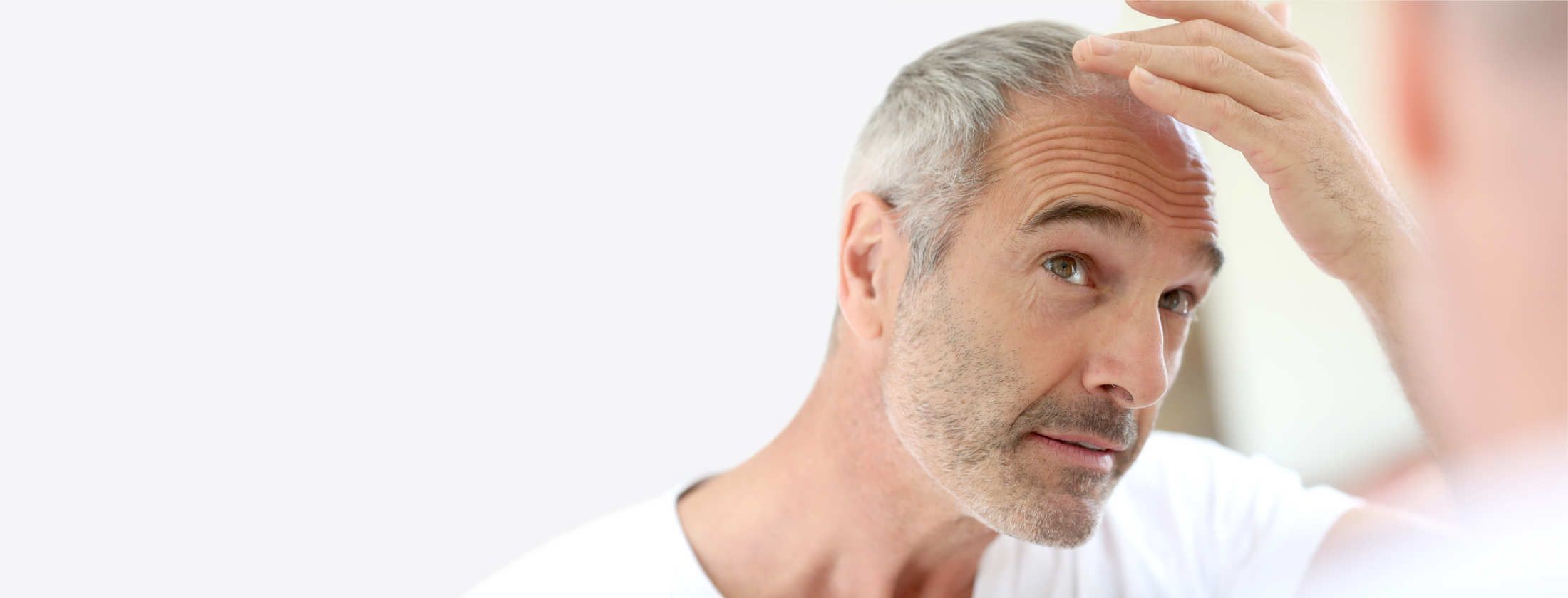 Grauhaariger Mann begutachtet sein rares Kopfhaar im Spiegel. Er ist in weißes Shirt gekleidet und denkt über eine Behandlung gegen Haarausfall nach. Anregung von Haarwachstum mit einem Haarwachstum-Serum. Mehr Haare würden seine Glatze verdecken.