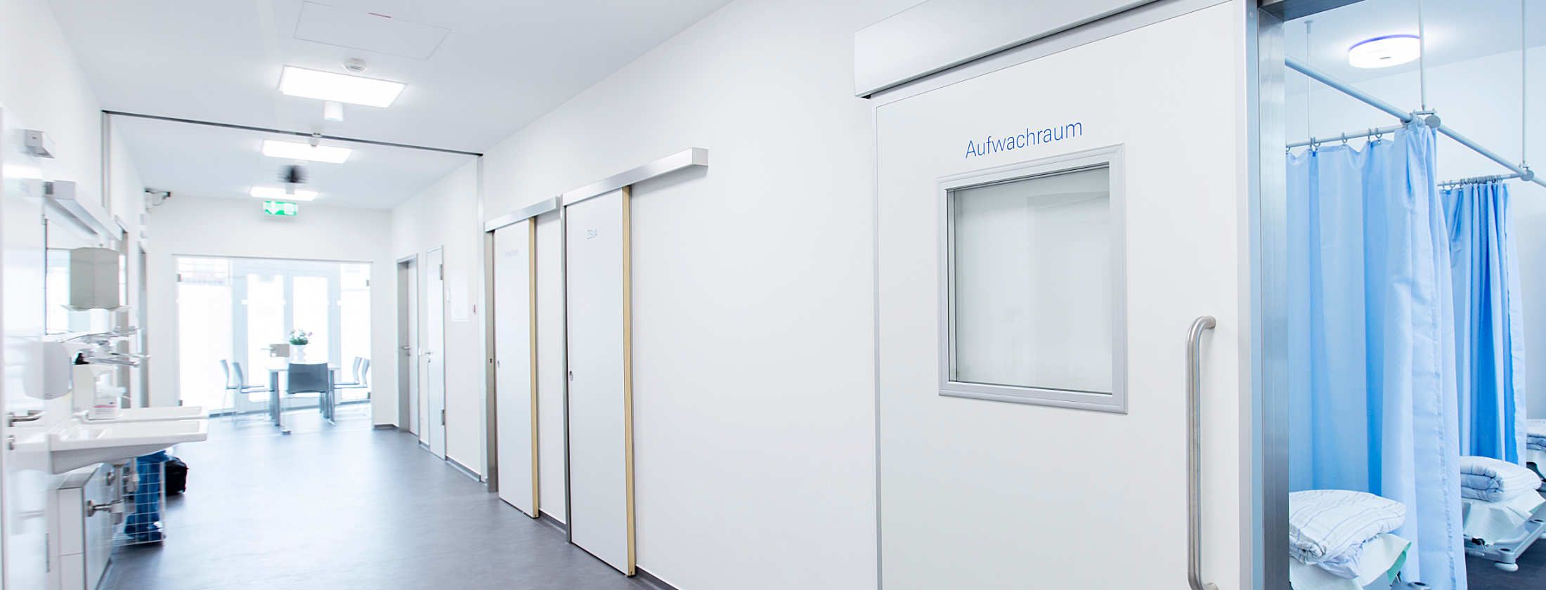 Langer heller Flur in der Berger Klinik Frankfurt. Türen sind an beiden Seiten. Flur der Privatklink endet mit wandgroßen Fenstern. Geöffnete Tür mit der Aufschrift Aufwachraum gewährt einen Blick in einen Raum voller Betten für ambulante Patienten.
