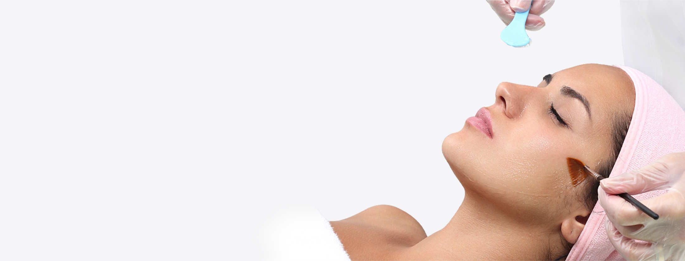 Frau mit einem gepflegten Gesicht liegt entspannt in ein Handtuch gehüllt. Eine Kosmetikerin trägt Fruchtsäure auf ihre Gesichtshaut auf. Die Flüssigkeit wirkt regenerierend auf die Haut. Anschließend wird per Peeling die oberste Hautschicht abgetragen.