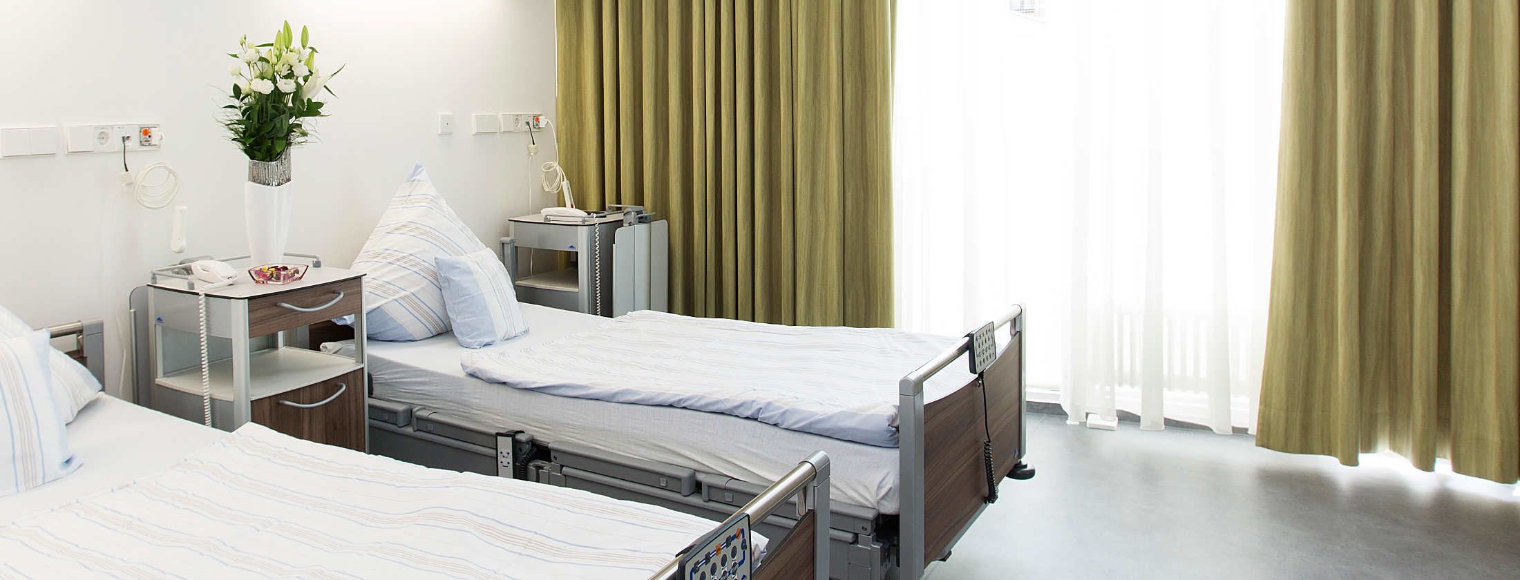 Patientenzimmer der Berger Klink in Frankfurt am Main für stationäre PatientInnen. Die Zimmer sind im sterilen Weiß gehalten. Die Gardinen im Goldton. Weiche Betten, Flachbildschirm und modernste Kommunikationsmittel bieten Komfort und Erholung nach OP.