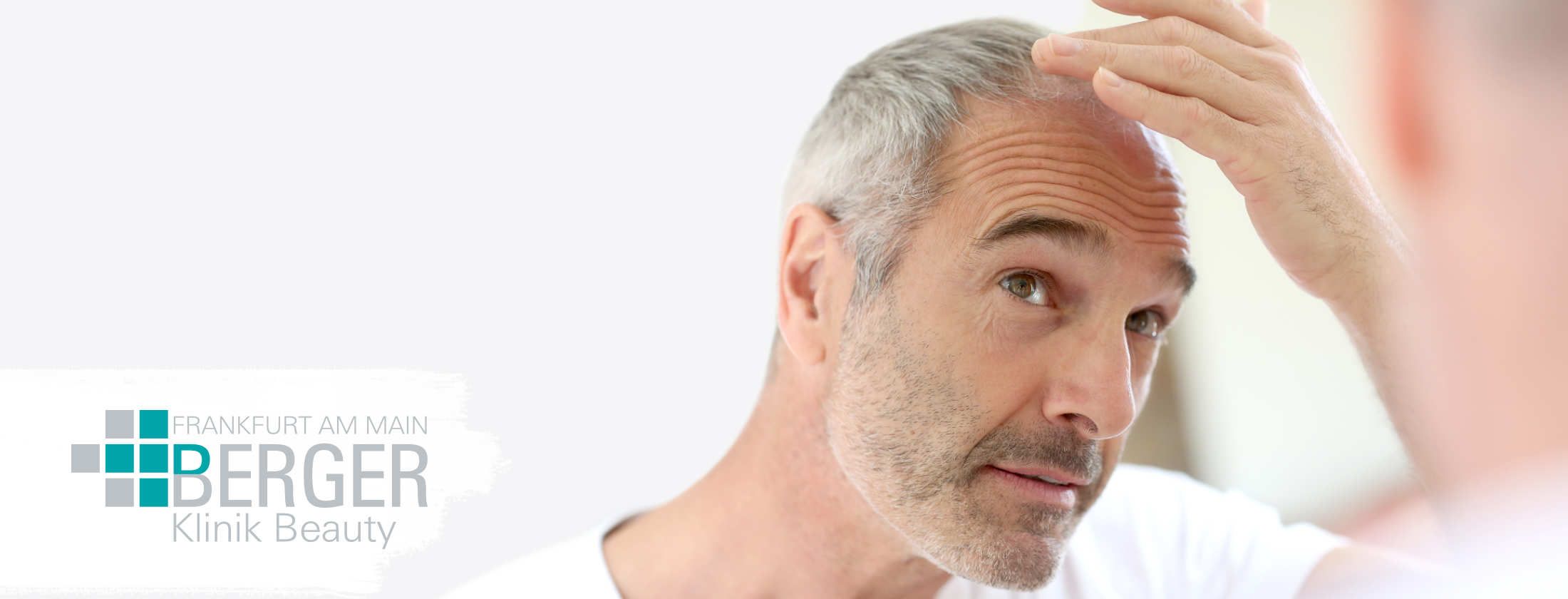 Grauhaariger Mann begutachtet sein rares Kopfhaar im Spiegel und denkt über eine kosmetische Behandlung zur Förderung von Haarwachstum. Enthaarung durch eine Laser-Haarentfernung kommt für ihn nicht in Frage. Er ist in ein weißes Shirt gekleidet.