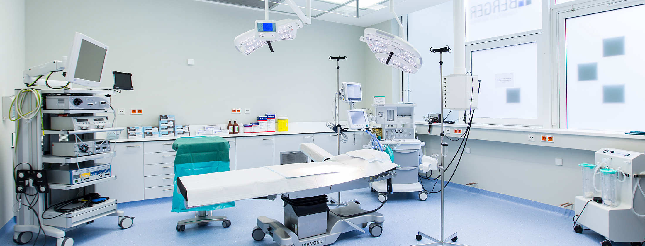 Operationssaal der Privatklinik im Frankfurter Westend. Mitten im Raum steht ein OP-Tisch, über ihn OP-Lampen. Im Hintergrund stehen Schränke mit Schubladen und Ablageflächen. Ein Anästhesiegerät zur Durchführung von Narkosen und ein Arthroskopieturm.