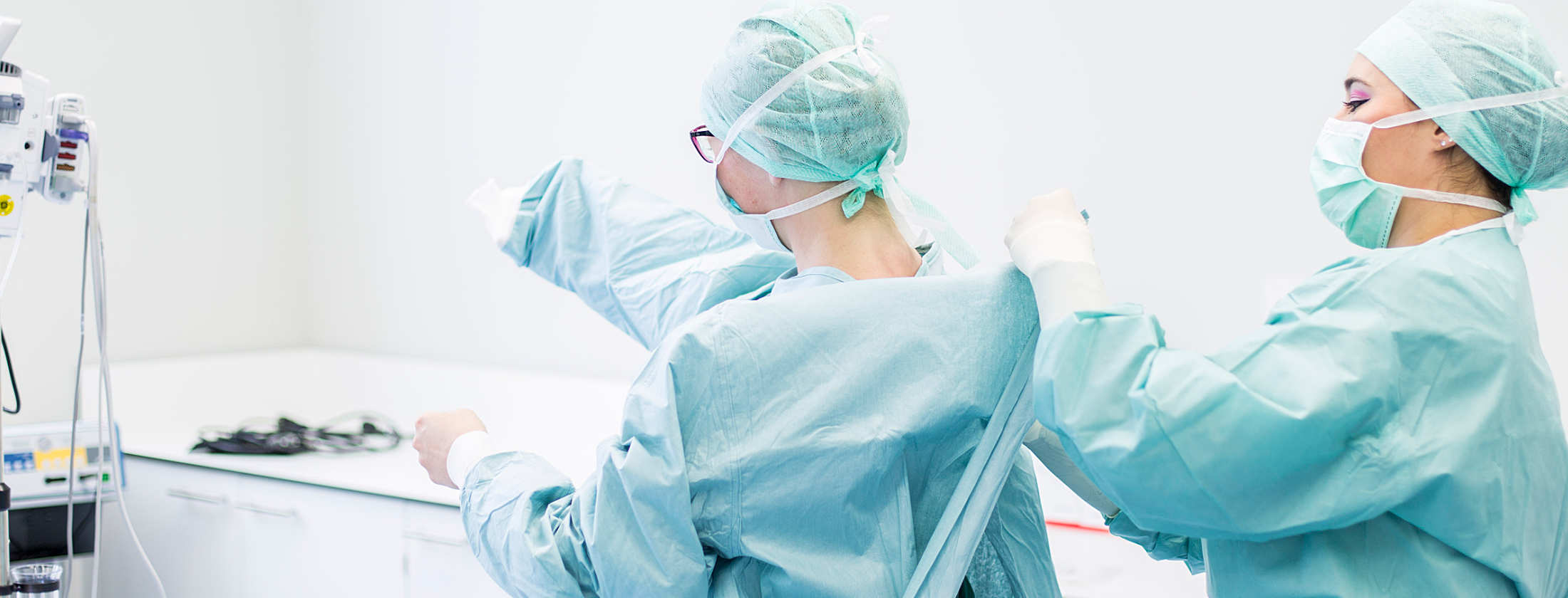 Fachärztin für Gynäkologie und Fachärztin für Urologie und Andrologie vor einem chirurgischen Eingriff. Vor der Operation kleiden sich die Ärztinnen in hygienisch saubere OP- Berufsbekleidung. Sie helfen sich gegenseitig die OP-Kittel überzuziehen.