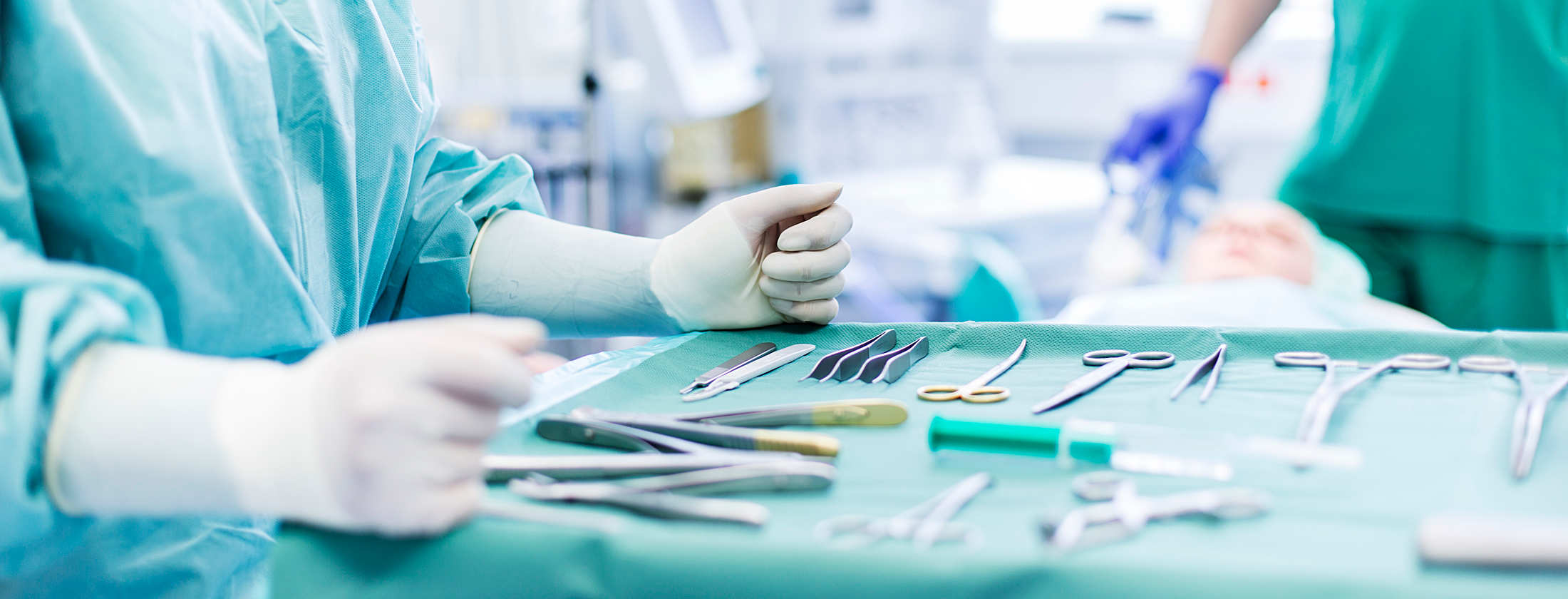 Operation eines Unfallpatienten in der Frankfurter Privatklinik. Im Hintergrund liegt eine betäubte Patientin. Im Vordergrund lehnt eine Medizinische Fachangestellte ihre Hände an einen OP-Tisch. Darauf sind viele chirurgische Instrumente zu sehen.